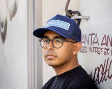 Load image into Gallery viewer, man wearing - blue suavecito hat with white embroidered text and light blue background &quot;suavecito&quot;
