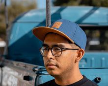 Load image into Gallery viewer, man wearing - blue suavecito hat with brown bill, brown text and brown background &quot;S suavecito&quot;
